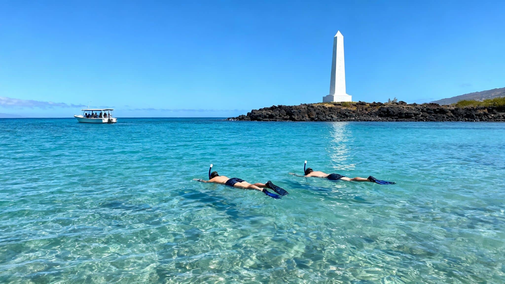 Two people snorkel in clear blue water near a white monument and a boat on a sunny day.