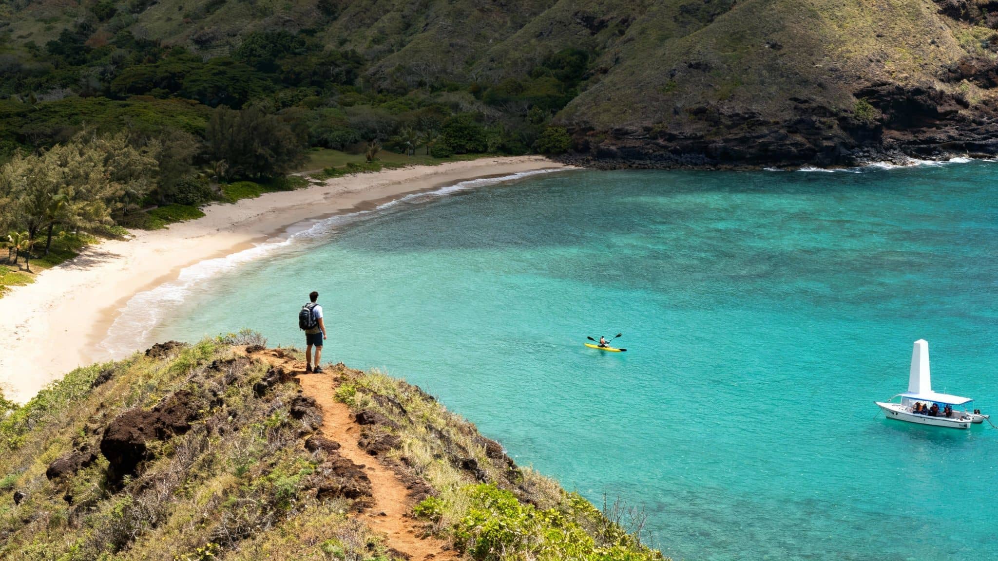 A hiker stands on a cliff overlooking a vibrant turquoise bay with a sandy beach, a kayak, and a boat.
