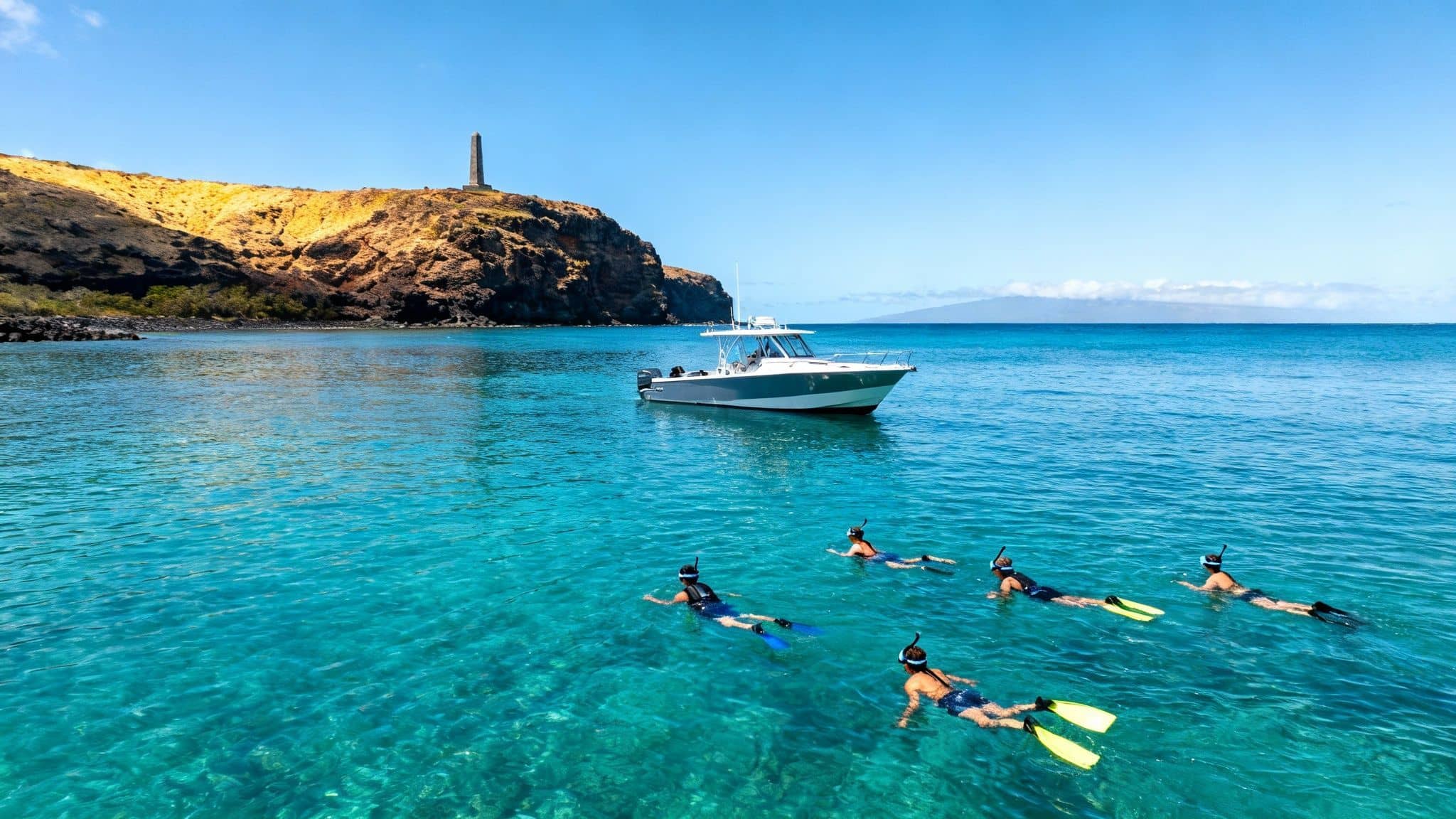 People snorkeling in clear blue water near a boat, a rocky island with a monument, and distant mountains.