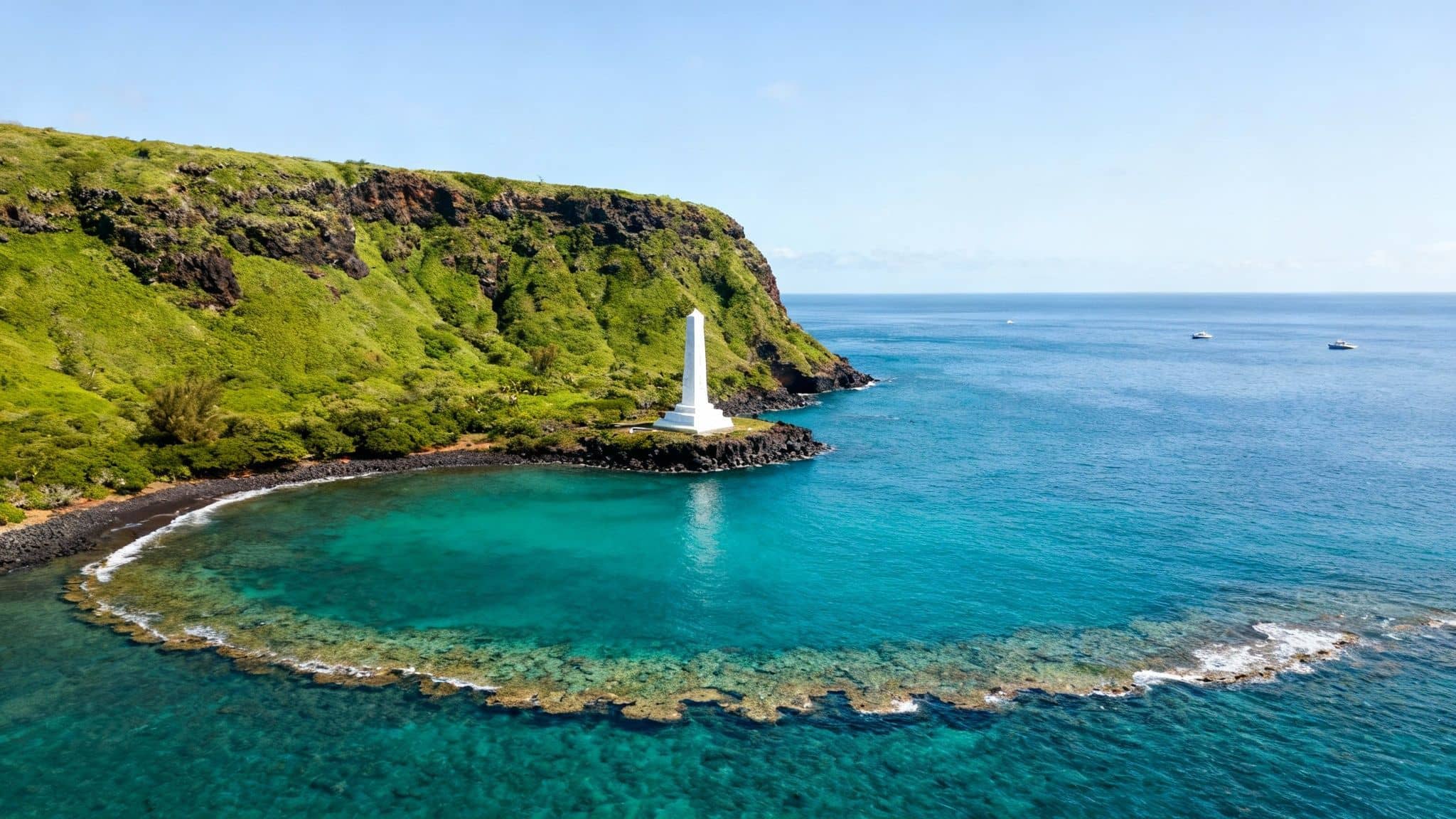 A panoramic view of the Kealakekua Bay Captain Cook Monument standing against a lush cliffside with turquoise water in the foreground.