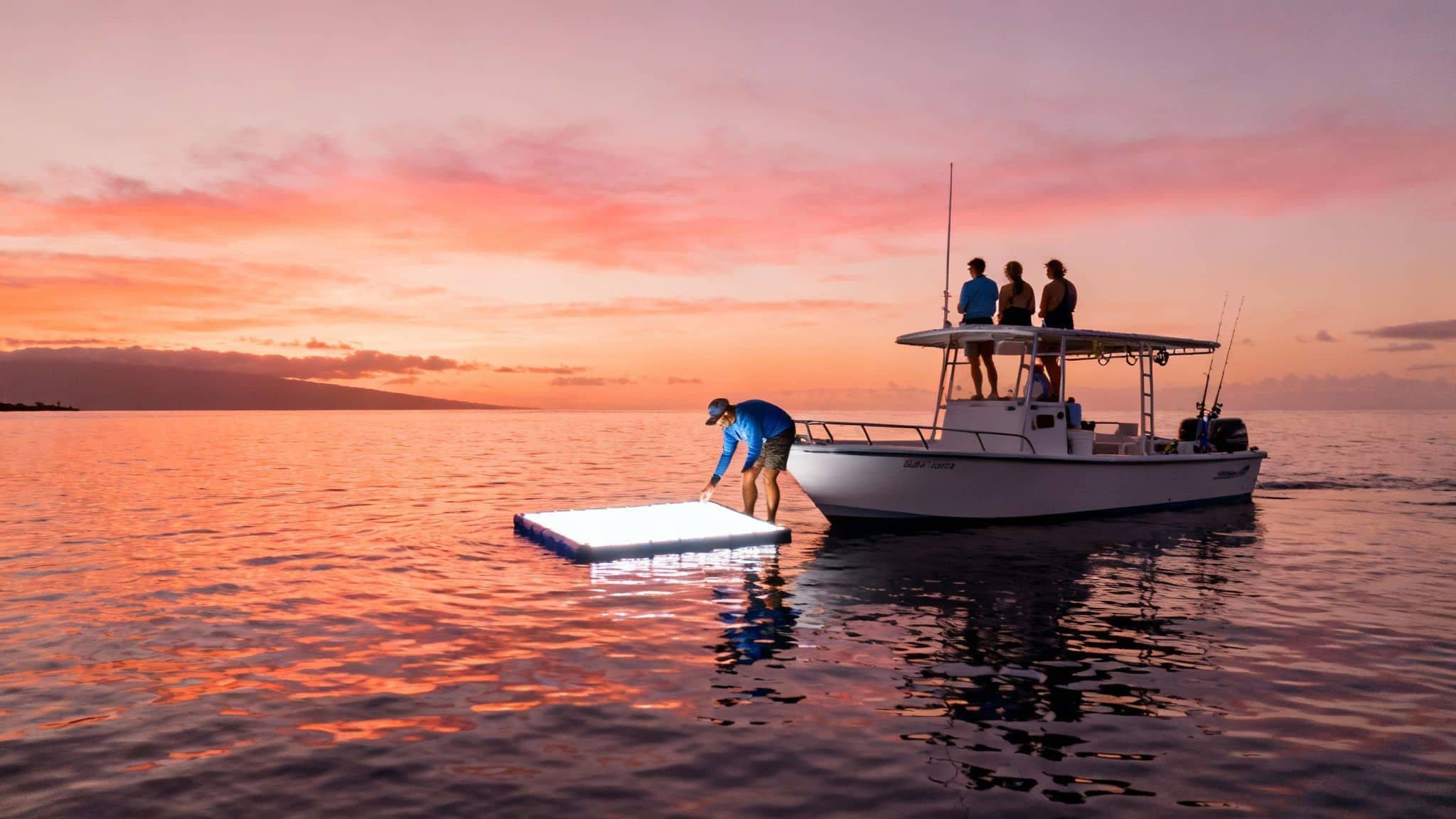 A man adjusts a large glowing platform in the ocean at sunset, near a boat with three people.