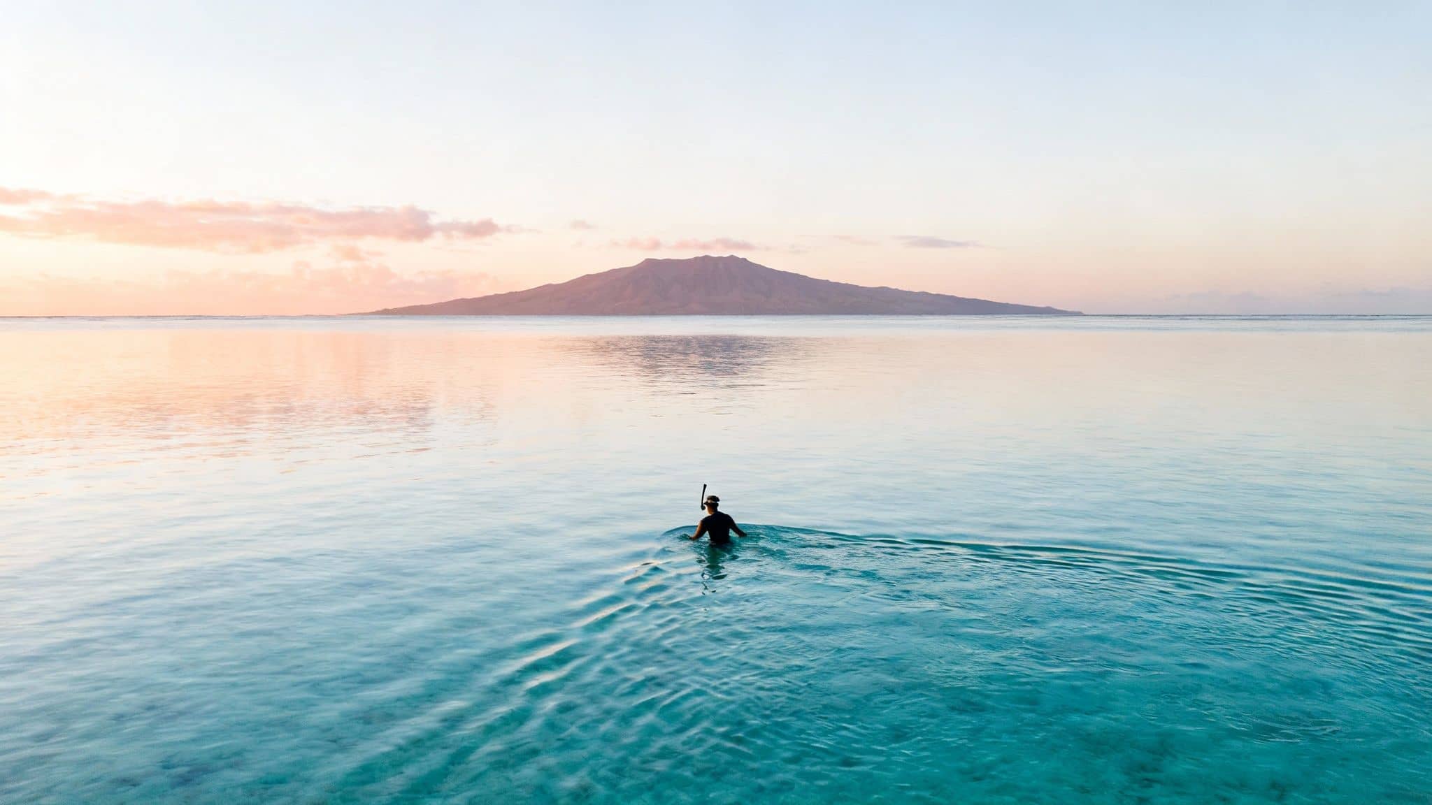 Person snorkeling in calm turquoise water with a distant island under a serene sunset sky.