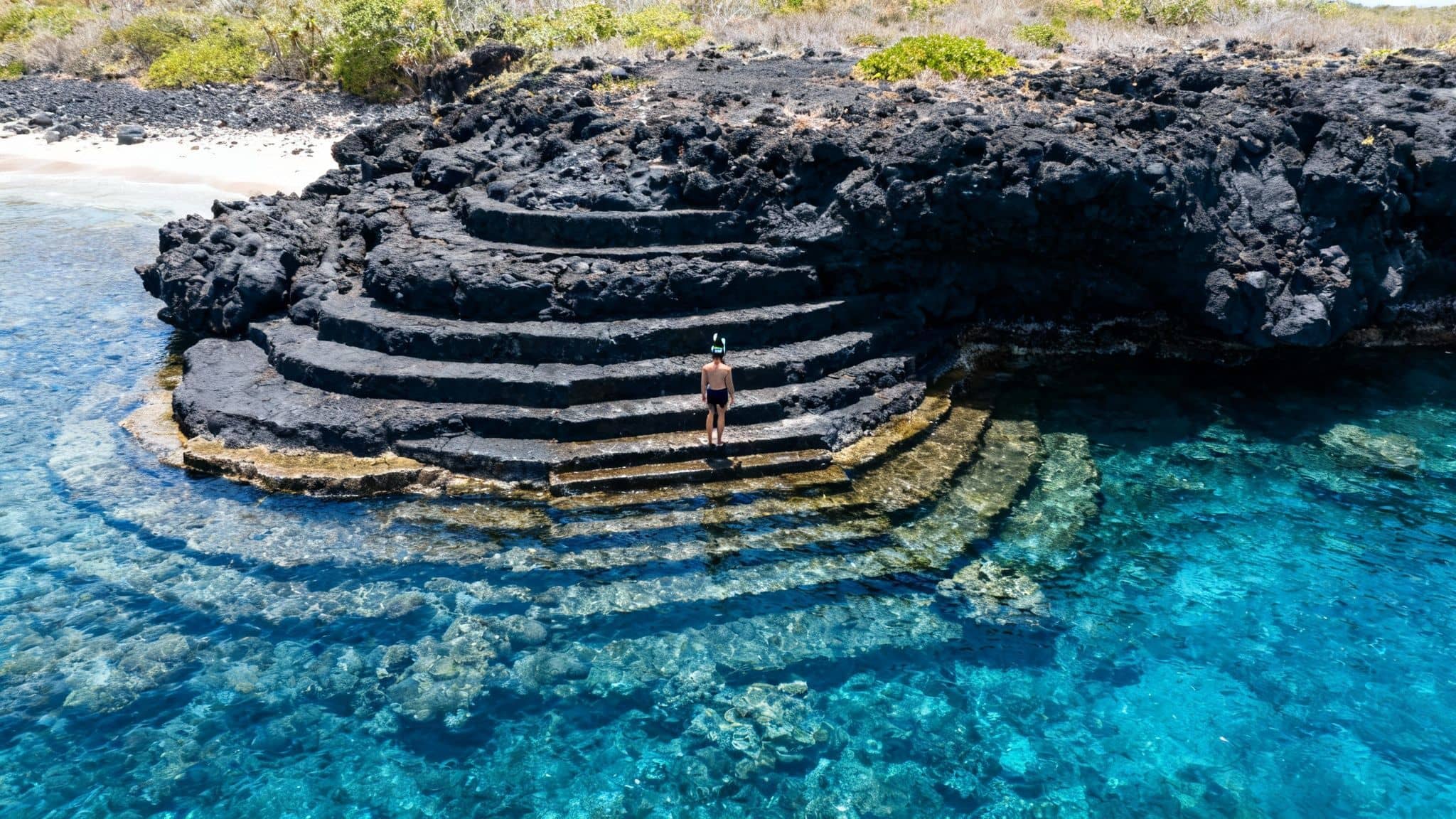Person with snorkel mask on dark volcanic steps leading into clear blue tropical ocean water.